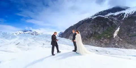 A bride and groom standing on a glacier during a wedding ceremony with mountains in the background.
