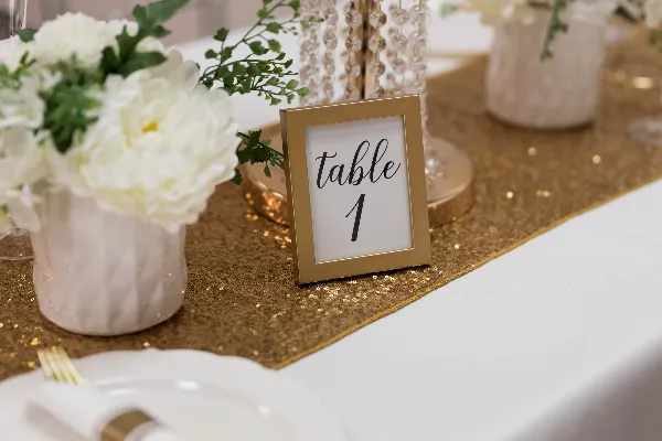 A close-up of a wedding table setting featuring a gold sequin runner, white floral arrangements, and a framed table number sign.
