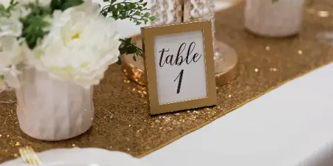A close-up of a wedding table setting featuring a gold sequin runner, white floral arrangements, and a framed table number sign.