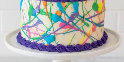 A round white cake with colorful abstract icing splatters and purple piped borders, displayed on a white cake stand.