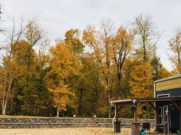 Outdoor view of The Wooded Garden venue featuring a stone wall, a building with a covered porch, and a backdrop of autumn trees.