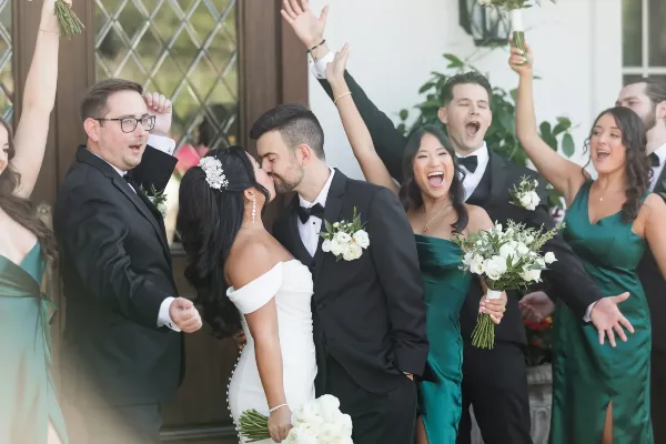 A bride and groom share a kiss while their wedding party cheers and raises their hands in celebration.