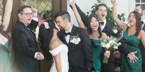 A bride and groom share a kiss while their wedding party cheers and raises their hands in celebration.