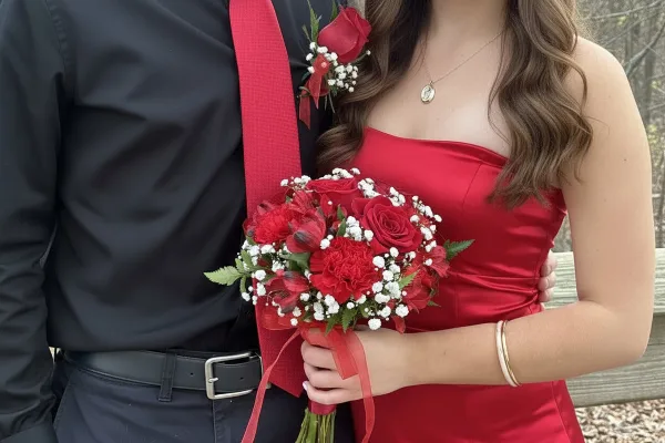 A person in a red dress holding a red rose and carnation bouquet with baby's breath, standing next to a person wearing a matching red rose boutonniere.