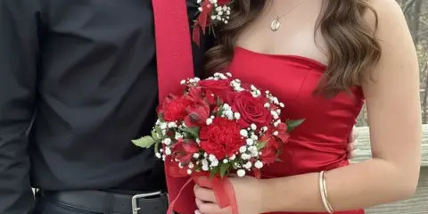 A person in a red dress holding a red rose and carnation bouquet with baby's breath, standing next to a person wearing a matching red rose boutonniere.