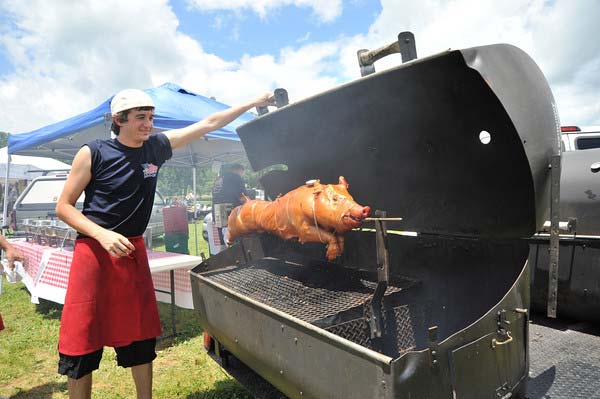 A person in a white cap and red apron tending to a whole pig roasting on a spit inside a large outdoor grill.