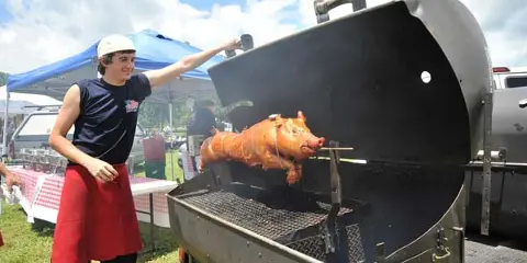 A person in a white cap and red apron tending to a whole pig roasting on a spit inside a large outdoor grill.