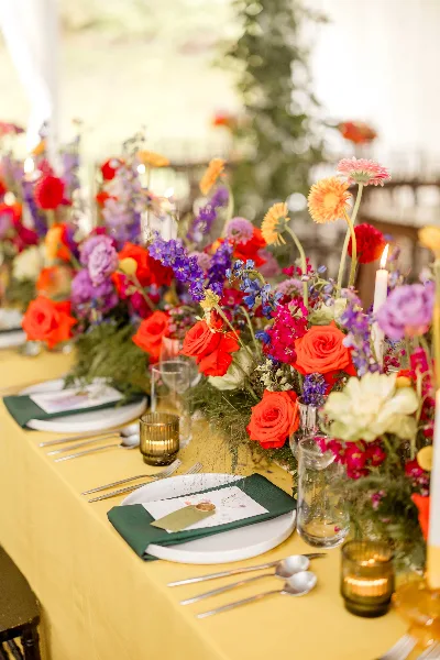 A vibrant, garden-style floral centerpiece featuring red roses, purple delphiniums, and orange gerbera daisies on a yellow tablecloth.