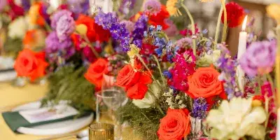 A vibrant, garden-style floral centerpiece featuring red roses, purple delphiniums, and orange gerbera daisies on a yellow tablecloth.