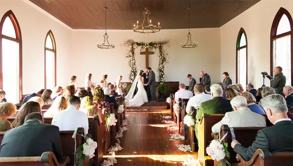A wedding ceremony in progress inside a chapel with a bride and groom at the altar and seated guests.