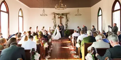 A wedding ceremony in progress inside a chapel with a bride and groom at the altar and seated guests.