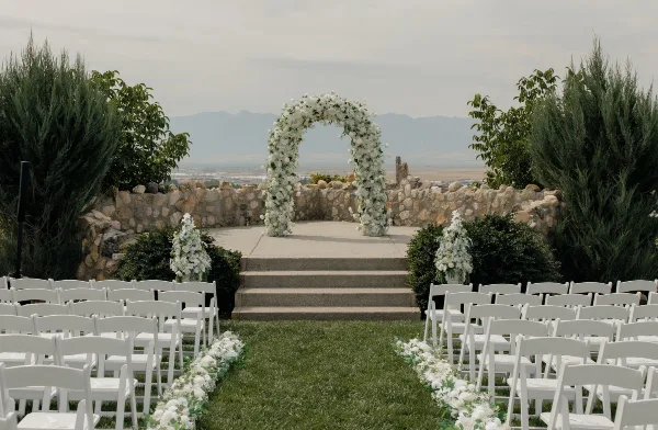 Outdoor wedding ceremony setup with a white floral arch, aisle flowers, and white chairs.