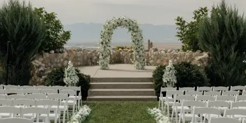 Outdoor wedding ceremony setup with a white floral arch, aisle flowers, and white chairs.