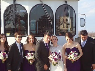 A wedding party including the bride and groom poses in front of a large white coach bus.