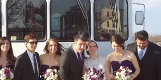 A wedding party including the bride and groom poses in front of a large white coach bus.