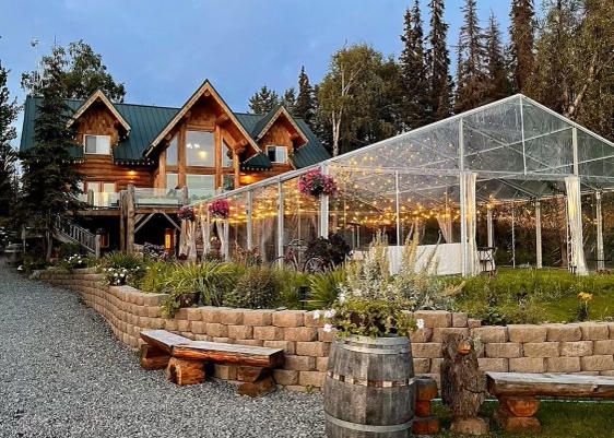 Exterior view of a log cabin lodge with a glass-walled event pavilion decorated with string lights and flowers.