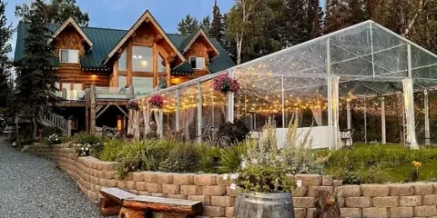 Exterior view of a log cabin lodge with a glass-walled event pavilion decorated with string lights and flowers.