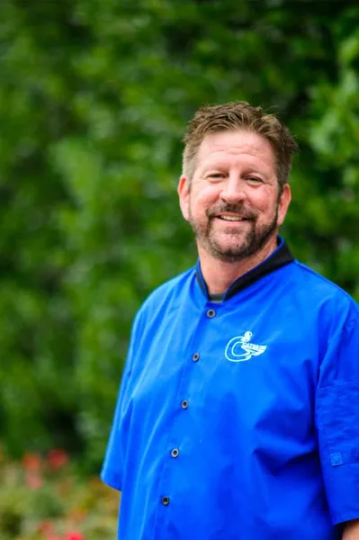 A smiling man in a blue chef's coat with a C2 Catering logo, standing outdoors in front of greenery.