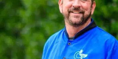 A smiling man in a blue chef's coat with a C2 Catering logo, standing outdoors in front of greenery.
