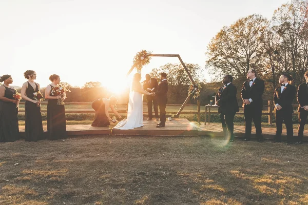 A bride and groom exchanging vows at a wooden arch during an outdoor wedding ceremony at sunset, with bridesmaids and groomsmen standing on either side.