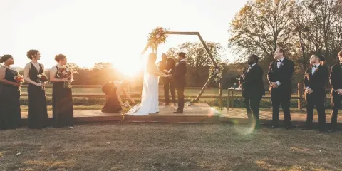 A bride and groom exchanging vows at a wooden arch during an outdoor wedding ceremony at sunset, with bridesmaids and groomsmen standing on either side.