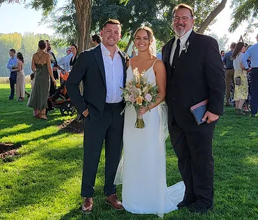A bride and groom standing with their officiant, Pastor Dave, outdoors after a wedding ceremony.