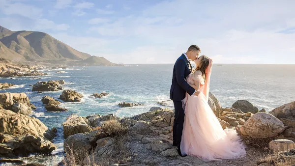 A bride and groom embracing on a rocky coastal cliff overlooking the ocean.