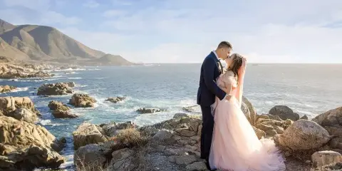 A bride and groom embracing on a rocky coastal cliff overlooking the ocean.