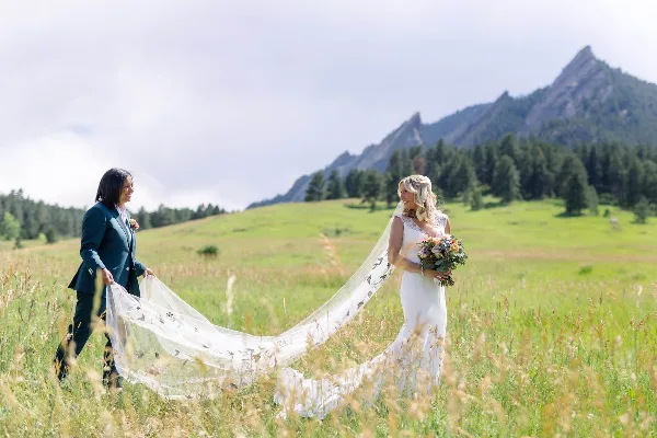 A bride in a white wedding dress and veil stands in a grassy field with a mountain in the background, holding a bouquet.