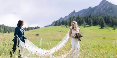 A bride in a white wedding dress and veil stands in a grassy field with a mountain in the background, holding a bouquet.