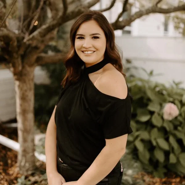 Smiling woman with long brown hair wearing a black off-shoulder top, standing outdoors near a tree and greenery.