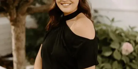 Smiling woman with long brown hair wearing a black off-shoulder top, standing outdoors near a tree and greenery.