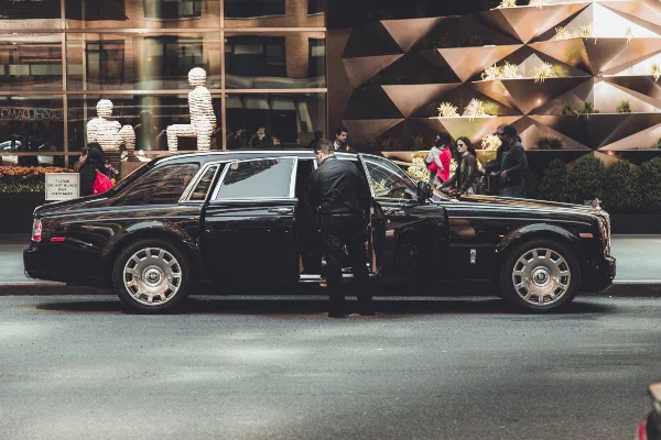 A black luxury limousine parked on a city street with a chauffeur standing by the open door.