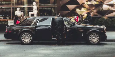 A black luxury limousine parked on a city street with a chauffeur standing by the open door.