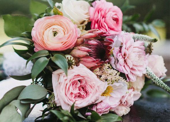 A close-up photograph of a floral bouquet featuring pink ranunculus, a protea flower, and greenery.
