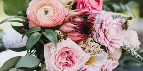 A close-up photograph of a floral bouquet featuring pink ranunculus, a protea flower, and greenery.
