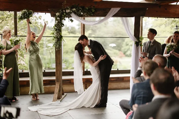 A bride and groom share a kiss under a floral arch during a wedding ceremony while guests and bridesmaids cheer.