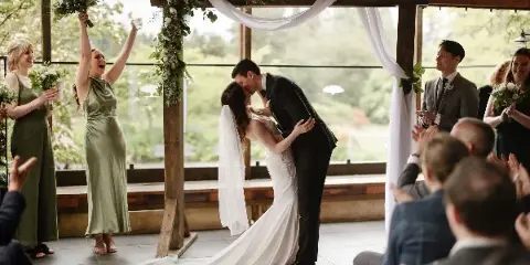 A bride and groom share a kiss under a floral arch during a wedding ceremony while guests and bridesmaids cheer.