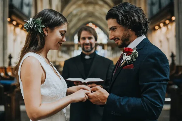 A bride and groom exchanging rings during a wedding ceremony with an officiant in the background.