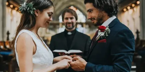 A bride and groom exchanging rings during a wedding ceremony with an officiant in the background.