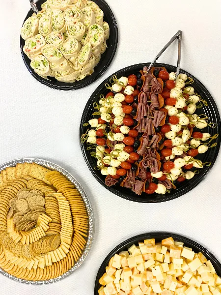 A catering spread featuring a charcuterie board with meats and cheese, a platter of tortilla pinwheels, and a tray of assorted crackers.