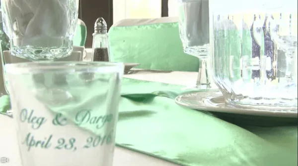 A close-up of a wedding table setting featuring a personalized glass with the names 'Oleg & Darya' and the date 'April 28, 2010', set against a green table runner and glassware.