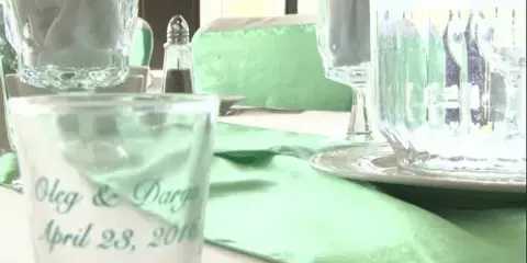 A close-up of a wedding table setting featuring a personalized glass with the names 'Oleg & Darya' and the date 'April 28, 2010', set against a green table runner and glassware.