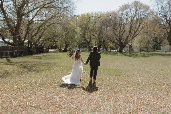A bride and groom holding hands and walking away from the camera in a grassy field with trees and string lights in the background.