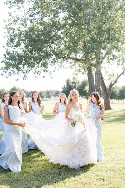 A bride in a white lace gown and her bridesmaids in light blue dresses posing outdoors on a lawn.