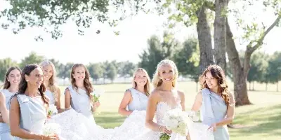 A bride in a white lace gown and her bridesmaids in light blue dresses posing outdoors on a lawn.