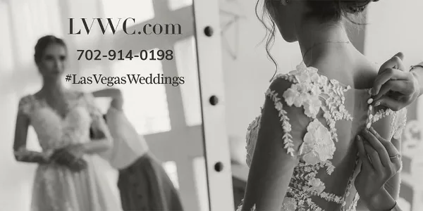 A bride in a floral lace wedding dress having her back buttoned by an assistant, with a mirror reflection visible in the background.