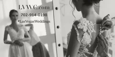 A bride in a floral lace wedding dress having her back buttoned by an assistant, with a mirror reflection visible in the background.