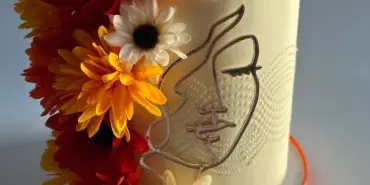 A white cake decorated with vibrant orange and yellow flowers and an artistic face design.