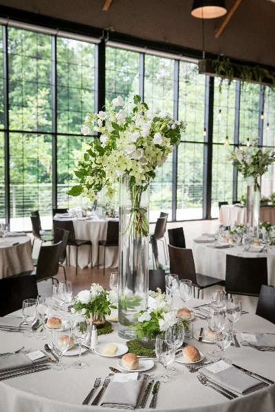 A tall, elegant floral arrangement with white flowers and greenery in a clear glass vase, placed on a set wedding reception table.
